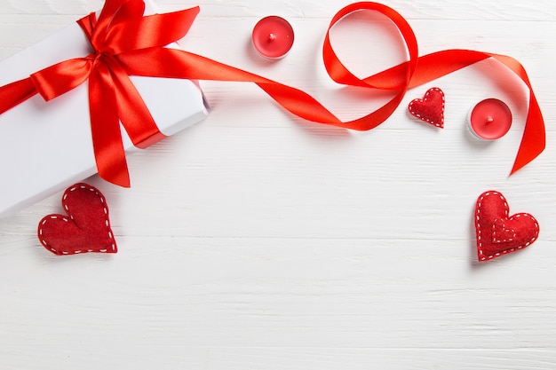 Regalo embalado blanco con cinta roja, velas y corazón hecho a mano sobre la mesa. Una romántica sorpresa de San Valentín para un ser querido.