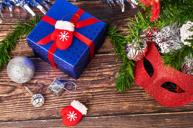 Un regalo en una envoltura azul, atado con una cinta roja, se encuentra sobre una mesa de madera, con oropel plateado, bolas navideñas y pequeñas manoplas rojas decorativas, una máscara de disfraces.