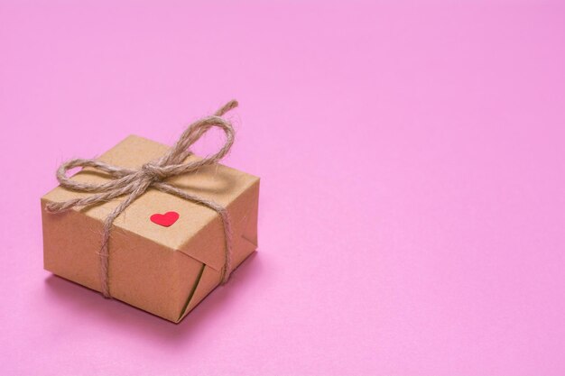 Un regalo envuelto en papel kraft sobre un fondo rosa Corazón rojo de papel en una caja de regalo