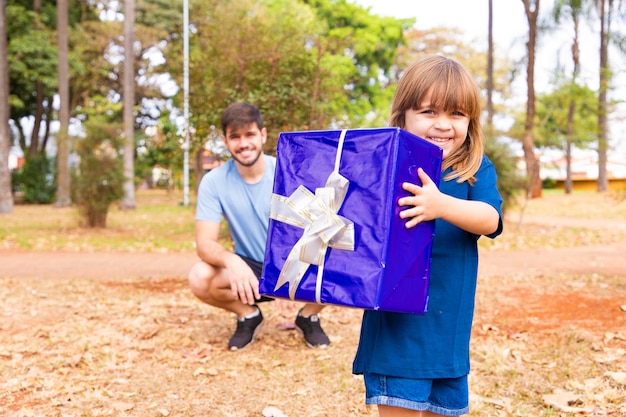 Este regalo es para ti retrato de una niña amorosa al lado del padre saludándolo con el día del padre o cumpleaños sosteniendo una caja de regalo envuelta felices vacaciones familiares juntos en el parque