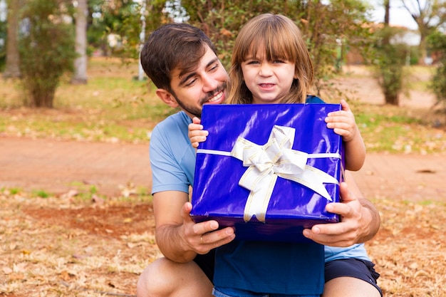 Este regalo es para ti Retrato de una niña amorosa al lado del padre saludándolo con el día del padre o cumpleaños sosteniendo una caja de regalo envuelta felices vacaciones familiares juntos en el parque