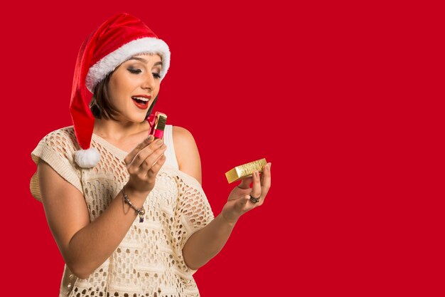 Regalo de Navidad - regalo de apertura de mujer sorprendido y feliz, joven y bella mujer sonriente con sombrero de Santa. Divertida foto linda de mujer morena sobre fondo rojo.