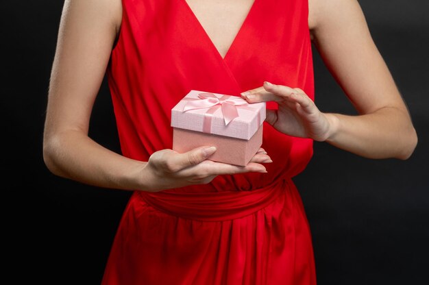 Regalo de vacaciones Sorpresa festiva Manos de mujer agraciada en vestido de cóctel rojo con caja de regalo rosa aislada sobre fondo oscuro Ocasión festiva Día especial