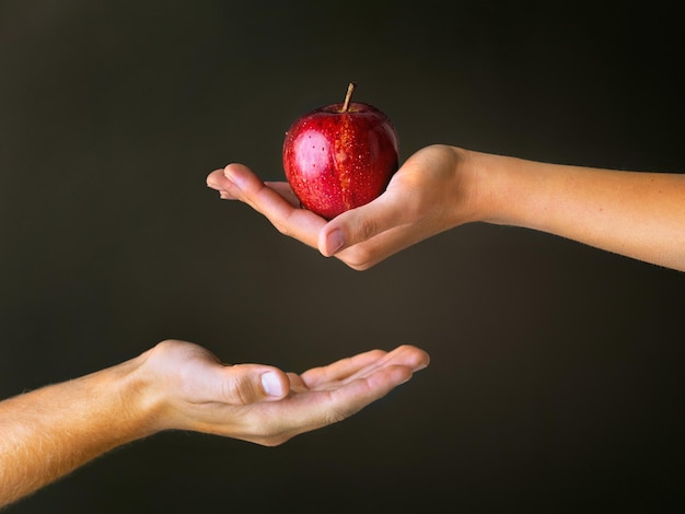 Regalos del Jardín del Edén Foto de estudio recortada de una mujer que ofrece a un hombre una manzana roja contra un fondo oscuro