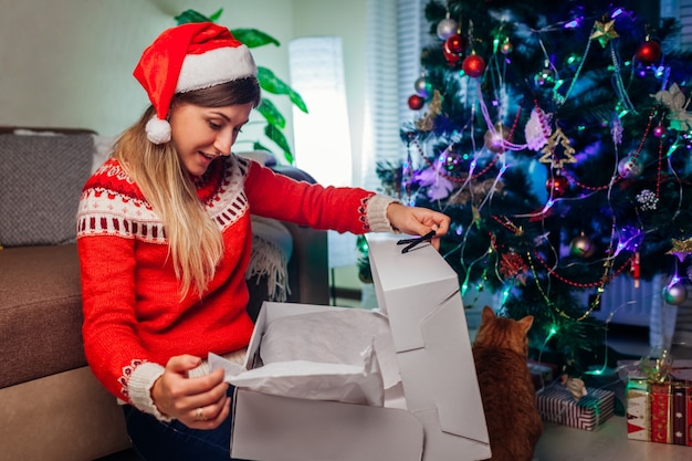 Regalos de Navidad y año nuevo Mujer con sombrero de Papá Noel abriendo caja de regalo por árbol de Navidad en casa