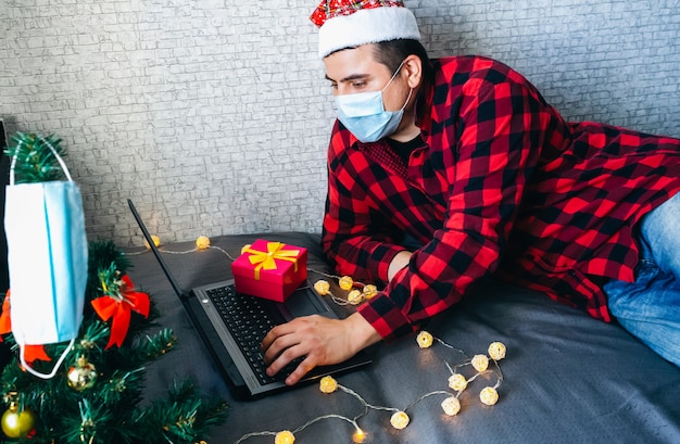 Regalos de Navidad dando. Hombre con máscara médica compartiendo regalos de caja a través de videollamadas. Vacaciones en aislamiento. Árbol de año nuevo y luces con adornos. Familiares y amigos celebrando en línea.