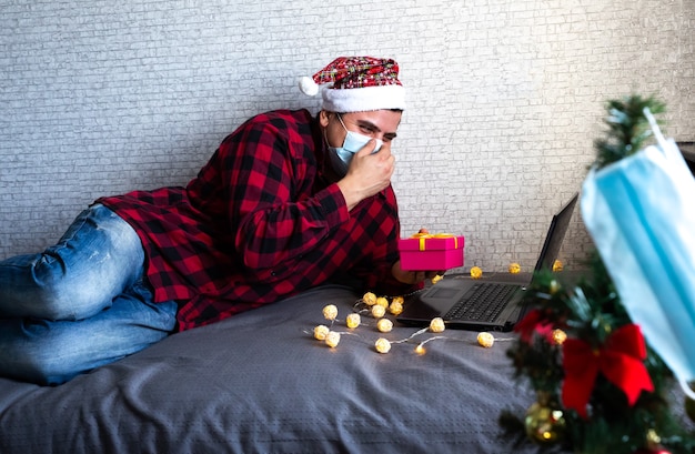 Regalos de Navidad dando. Hombre con máscara médica compartiendo regalos de caja a través de videollamadas. Vacaciones en aislamiento. Árbol de año nuevo y luces con adornos. Familiares y amigos celebrando en línea.