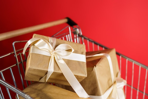 Regalos en papel artesanal con cinta blanca en un carro de supermercado sobre un fondo rojo.