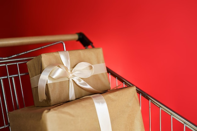 Regalos en papel artesanal con cinta blanca en un carro de supermercado sobre un fondo rojo.