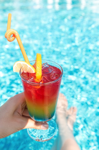 Regenbogencocktail mit Eis in weiblicher Hand durch den Pool.