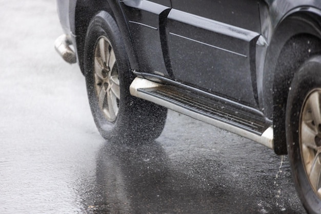Regenwasserspritzer fließen von den Rädern eines schwarzen Autos, das sich schnell in der Tageslichtstadt mit selektivem Fokus bewegt
