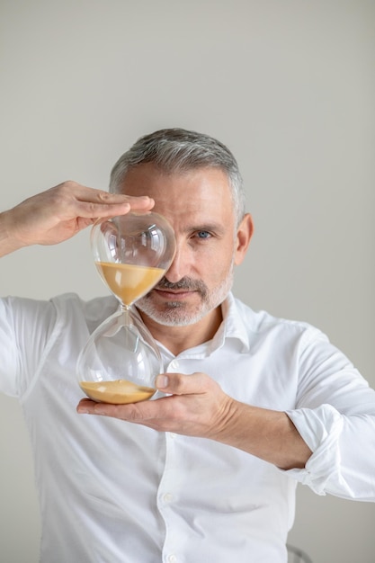 Regla importante. Exitoso hombre de mediana edad de cabello gris mirando a la cámara sosteniendo un reloj de arena al nivel de la cara de pie contra un fondo claro