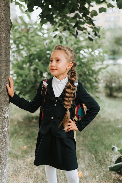 De regreso a la escuela, niña feliz, alumna, colegiala de ocho años con uniforme de moda con mochila y peinado, voluminosa trenza larga lista para ir al segundo grado, primer día en la escuela primaria