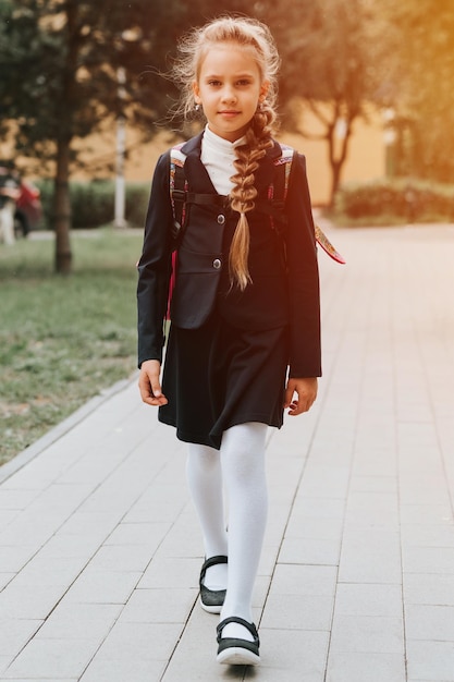 Regreso a la escuela, niña feliz, alumna, colegiala de ocho años con uniforme de moda con mochila y peinado, voluminosa trenza larga lista para ir al segundo grado, primer día en la escuela primaria