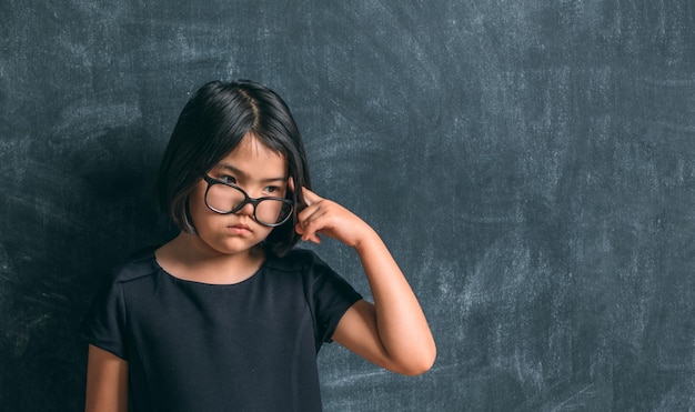 Regreso a la escuela Niña seria con gafas pensando en tocarse la cabeza con un dedo cerca de la pizarra Niño de la escuela primaria Educación