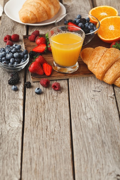 Reichhaltiger, leckerer Frühstückshintergrund. Croissants mit goldener Kruste, Glas frischem Orangensaft und vielen süßen Bio-Beeren auf Holzbrett über rustikaler Oberfläche. Köstlicher Start in den Tag, Kopierraum