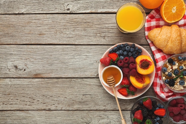 Reichhaltiger, leckerer Frühstückshintergrund. Croissants mit goldener Kruste, Müsli, Honig, Glas frischem Orangensaft und Schüssel mit süßen Bio-Heidelbeeren auf rustikaler Holzoberfläche, Draufsicht, Kopierraum