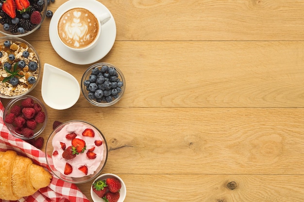 Reichhaltiges kontinentales Frühstück im Hintergrund. Croissants mit französischer Kruste, Müsli, Joghurt, süße Beeren und heißer Kaffee für leckere Mahlzeiten am Morgen. Köstlicher Start in den Tag. Ansicht von oben, kopieren Sie Platz auf Naturholz