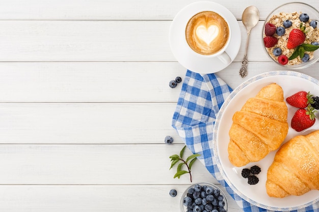 Reichhaltiges kontinentales Frühstück im Hintergrund. Knusprige französische Croissants, Müsli, viele süße Beeren und heißer Kaffee für leckere Morgenmahlzeiten. Köstlicher Start in den Tag. Ansicht von oben, kopieren Sie Platz auf weißem Holz