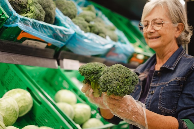 Foto reife ältere frau im supermarkt, die lebensmittel einkaufen und grünes brokkoliegemüse auswählt