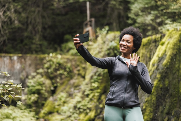 Reife Afroamerikanerin macht Selfie, während sie in der Nähe des Wasserfalls steht und die Aussicht während ihrer Wanderung in den Bergen genießt.