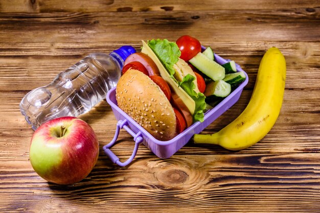 Reife Apfelbananenflasche Wasser und Brotdose mit Hamburgergurken und Tomaten auf Holztisch