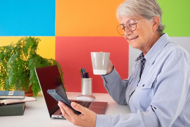 Reife attraktive Seniorin, die eine Kaffeetasse hält und Smartphone vom Büro aus verwendet Geschäftsfrau, die zu Hause arbeitet Online-Arbeitsinternet-Banking-Wireless-Technologie, die von zu Hause aus arbeitet