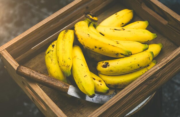 Foto reife bananen in einem holzbehälter und ein bananenmesser in natürlichem licht und dunkelgrauen tönen