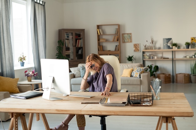 Reife blonde fröhliche Frau, die vor Computerbildschirm lacht, während sie am Tisch vor dem Monitor sitzt und Online-Film zu Hause sieht