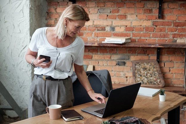 Reife Dame mit Brille, die am Hals hängt, der am Schreibtisch steht und Online-Dokument auf Laptop speichert