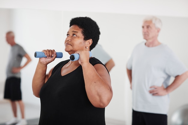 Reife dicke Frau, die Hanteln hebt, während sie zusammen mit anderen Leuten im Fitnessstudio trainiert