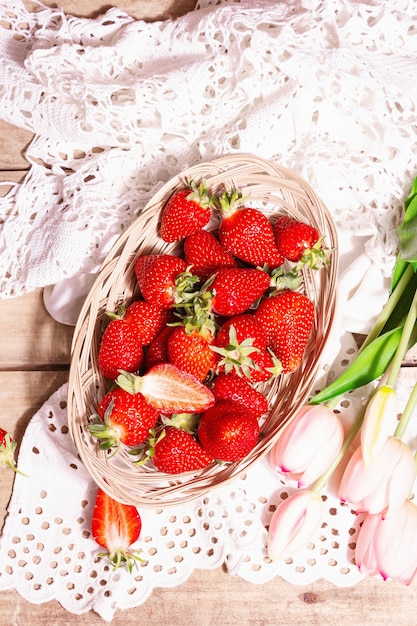 Reife Erdbeeren im Korbteller. Frische Beeren, aromatische Minze, rosa Tulpen. Alter Holztisch, Vintage gehäkelte Serviette, rustikaler Stil, Draufsicht