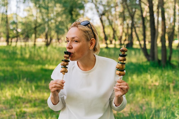 Reife Frau 50 Jahre alt, die im Sommerwald gegrilltes Grillgemüse von Holzspießen im Freien isst