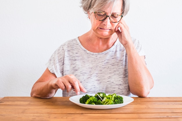 Reife Frau am Tisch, die mit ekelhaftem Gesicht auf gesundes Essen oder Brokkoli schaut - das Gemüse berührt und den Diät-Lebensstil nicht mag