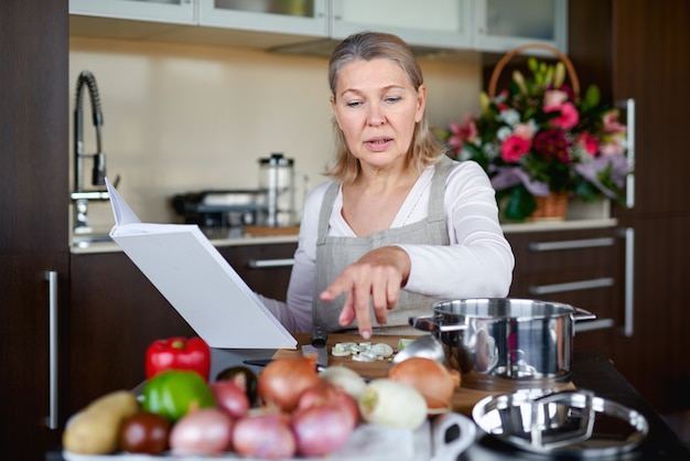 Foto reife frau, die essen in der küche vorbereitet und rezeptbuch betrachtet