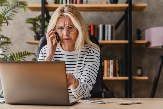 Reife Frau, die gestresst aussieht, während sie einen Laptop benutzt und mit dem Handy telefoniert