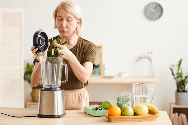 Foto reife frau, die gesunden cocktail aus spinat und früchten unter verwendung des mixers beim stehen in der küche macht