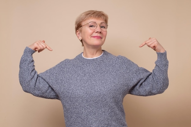 Reife Frau, die selbstbewusst mit einem Lächeln im Gesicht aussieht und sich mit stolzen und glücklichen Fingern zeigt