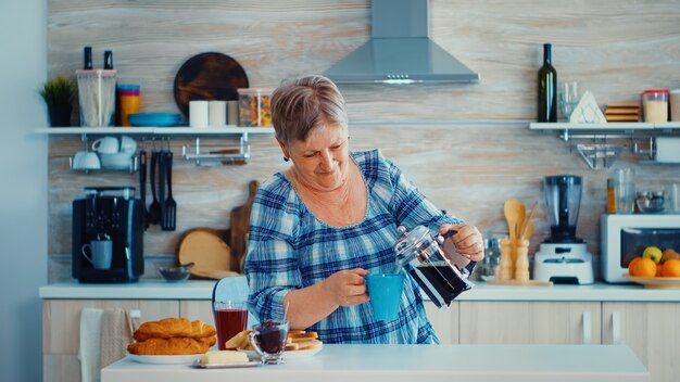 Reife Frau, die während des Frühstücks französische Presse verwendet, um Kaffee in der Küche zuzubereiten. Ältere Menschen genießen morgens frisches braunes Kaffee-Espresso-Koffein aus Vintage-Becher, Filter entspannen Erfrischung