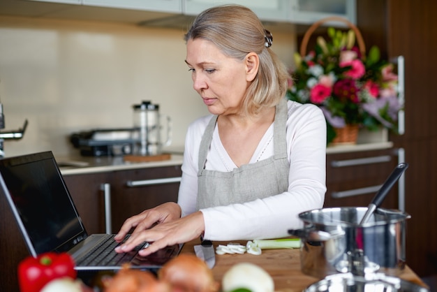 Reife Frau in der Küche bereitet Essen zu und schaut in Laptop