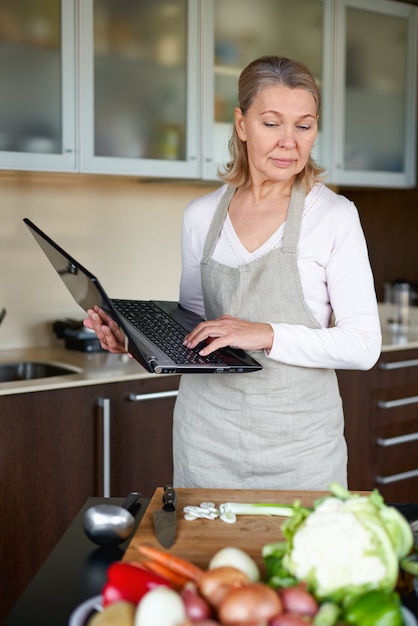 Reife Frau in der Küche, die Essen vorbereitet und Laptop hält