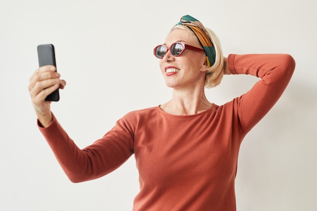 Reife Frau in der Sonnenbrille, die selfie auf ihrem Handy gegen den weißen Hintergrund macht