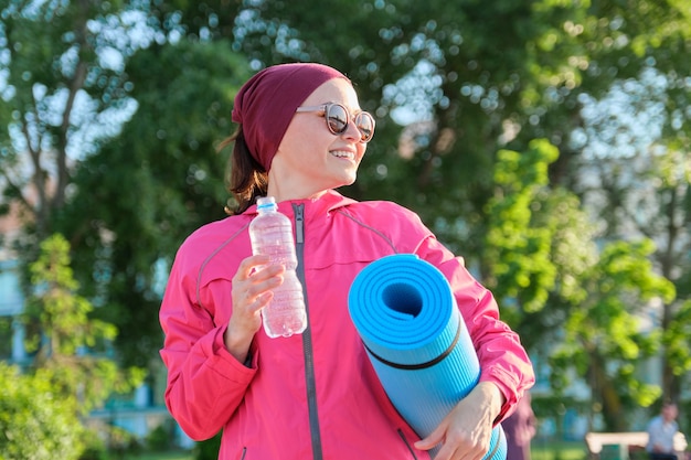 Reife Frau in Sportwindjacke mit Yogamatte Flasche Wasser, Wandern im Freien. Aktiver gesunder Lebensstil, Sport, Fitness bei Menschen mittleren Alters