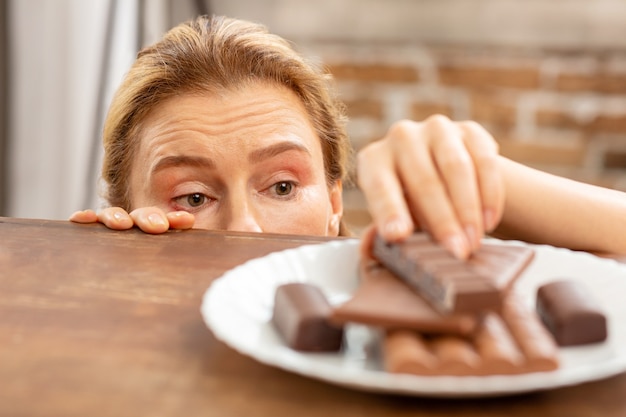 Reife Frau mit Allergie, die ein Stück Schokolade vom Tisch nimmt