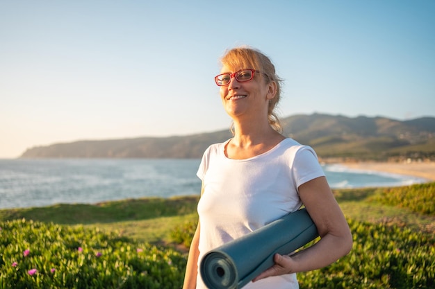 Foto reife frau mit brille hält eine yoga-matte und steht am strand