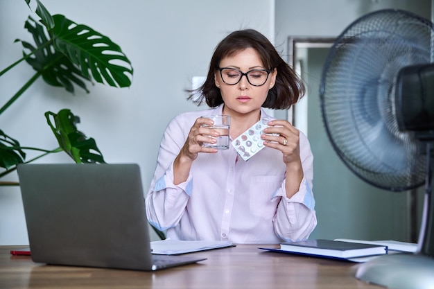 Reife Frau mit einem Glas Wasser, einer Blase voller Pillen, die Medikamente und Vitamine zur Linderung der Wechseljahrsbeschwerden einnimmt, sitzt am Schreibtisch am Arbeitsplatz mit einem Computer-Laptop