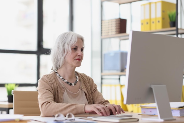 Reife Frau mit grauem Haar schaut auf den Bildschirm des Computermonitors und tippt auf der weiblichen Tastatur