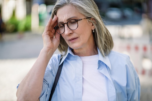 Reife Frau mit grauen Haaren in den 50ern und 60ern, die draußen steht und ihren Kopf hält, leidet unter Migränekopfschmerzen. Sie leidet unter Sommerzittern und fühlt sich während der Reise krank. Reife Frau steht draußen