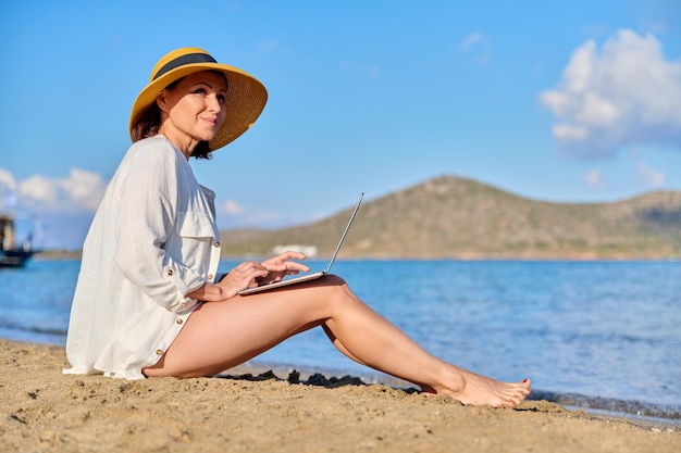 Reife Frau mit Hut mit Laptop am Meeresstrand