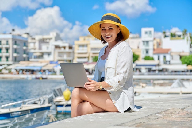 Reife Frau mit Hut und Laptop an der Strandpromenade in der Stadt